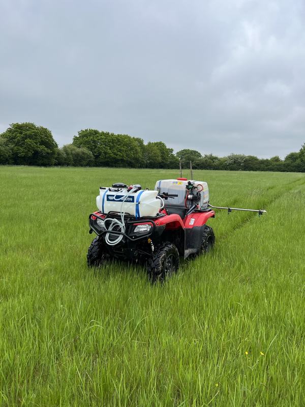 Red quad bike carrying spray tanks with a side boom, parked in a long-grass field