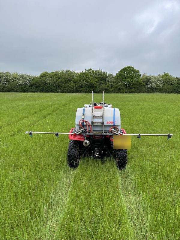 Red quad bike fitted with a tank and wide boom sprayer, spraying a grass field