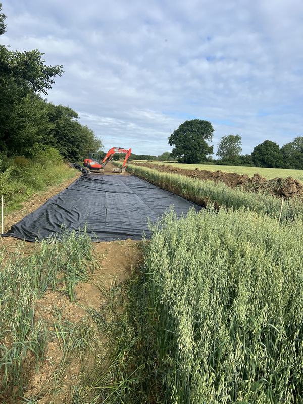Black geotextile membrane laid along the excavated track beside a crop field, with a digger in the distance