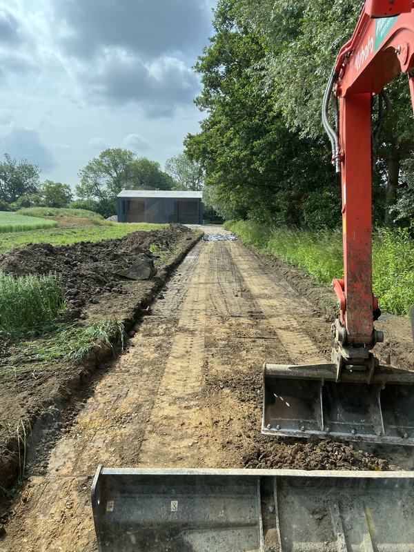 Farm access track stripped and excavated towards a barn, seen from the digger with the bucket in view
