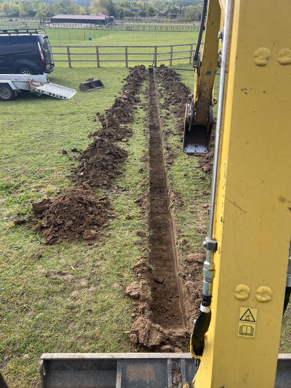 Narrow trench being dug across a grass paddock, seen from the mini digger cab