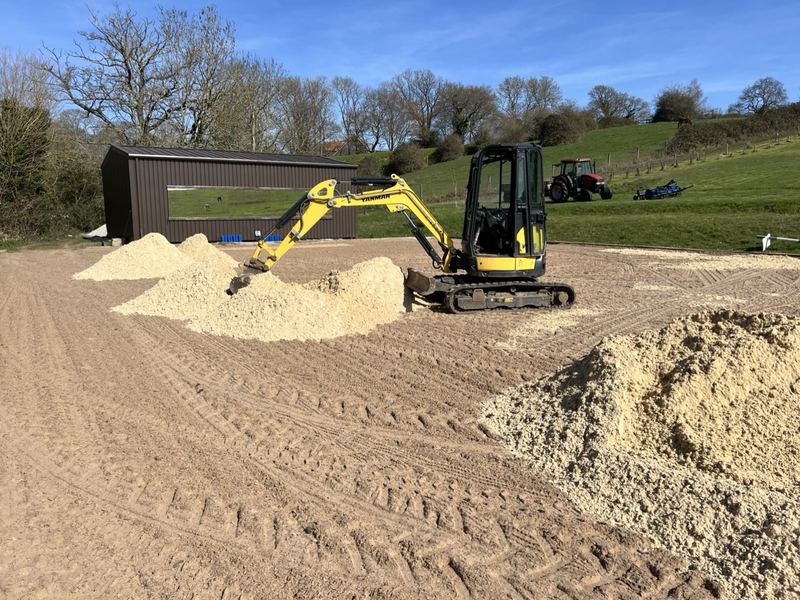 Yellow mini digger spreading heaps of new waxed silica sand mix across an outdoor ménage, with a timber stable block and a tractor behind
