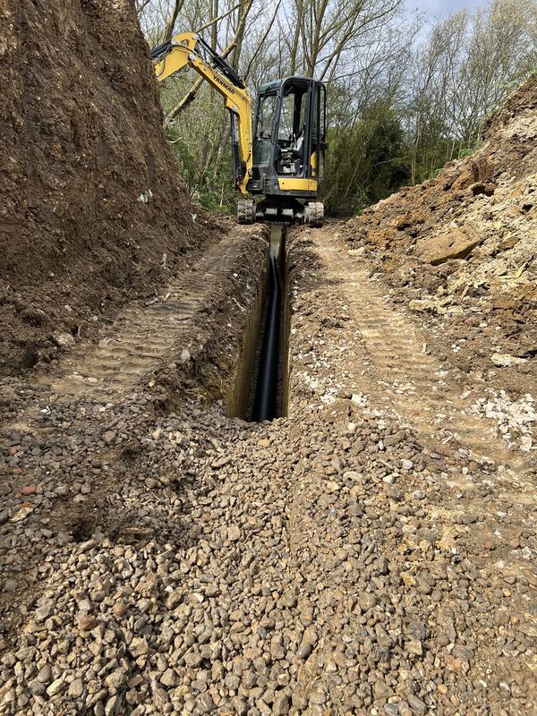 Yellow Yanmar mini digger laying land drain in a deep trench with stone backfill in a wooded area