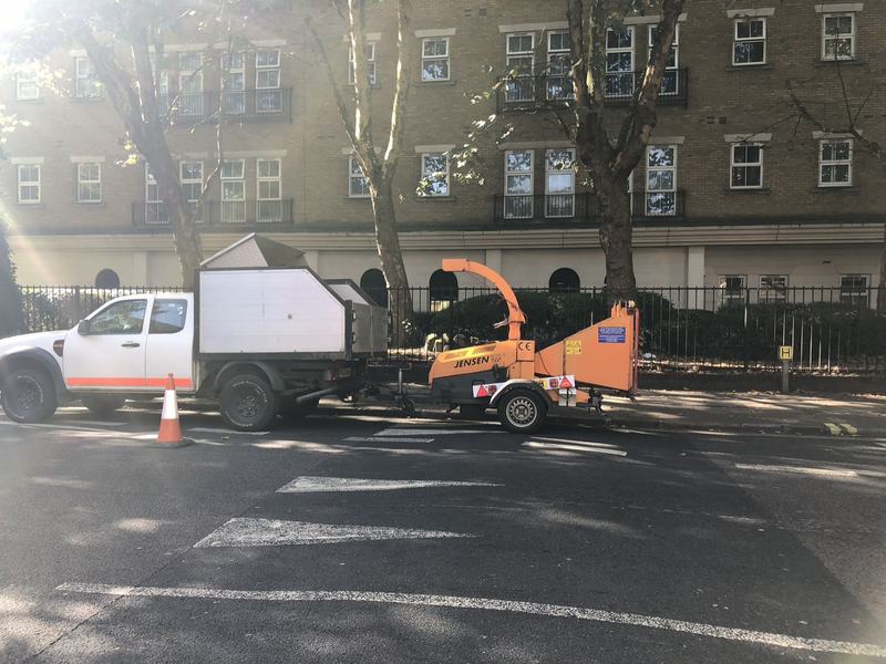 Pickup truck with a tipper body towing a Jensen wood chipper, parked beside a tree-lined pavement