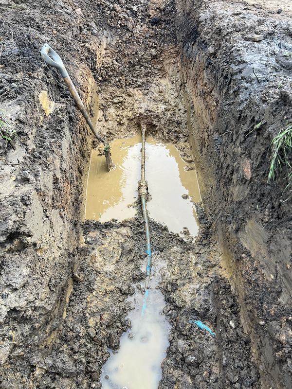 Deep trench excavated to expose a blue water supply pipe, with leaking water pooling in the bottom